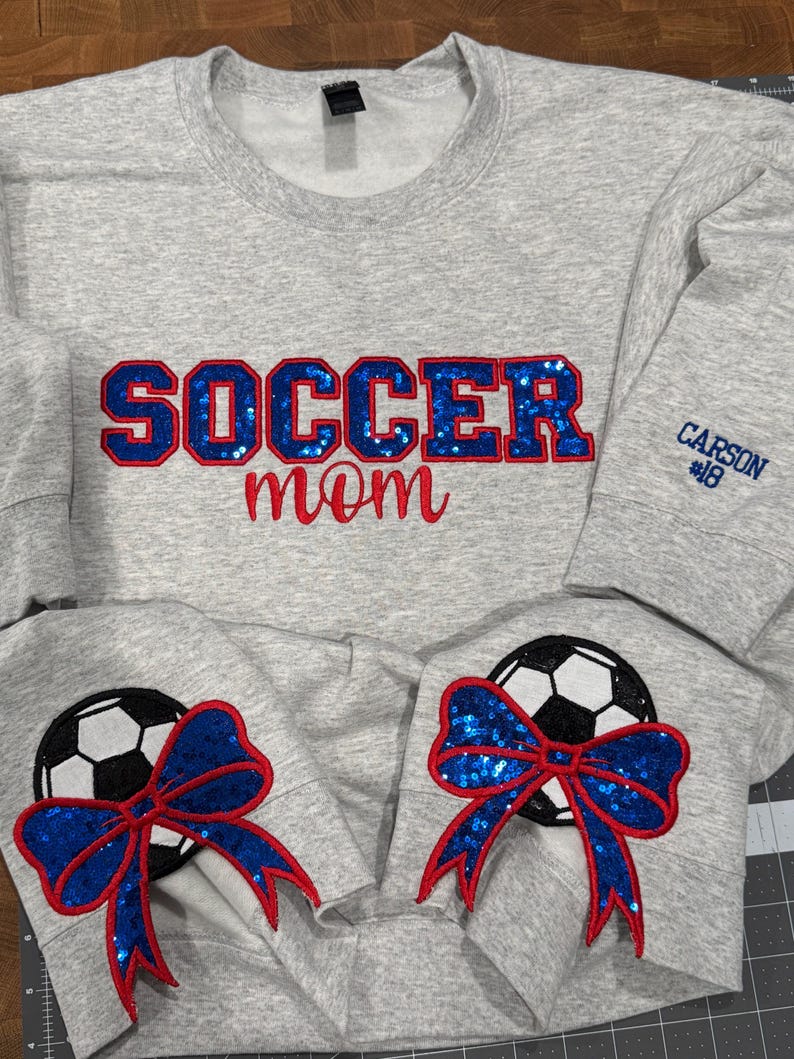 May include: A light gray crewneck sweatshirt featuring the text SOCCER in blue sequin letters with red outlines and mom in red script. The sleeves display soccer ball graphics with blue sequin bows and red outlines. CARSON #18 is embroidered in blue.