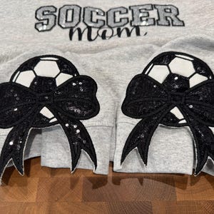 May include: A light gray sweatshirt featuring the text SOCCER MOM in black sequined letters. Each sleeve cuff displays a large black sequined bow layered over a white and black soccer ball graphic, creating a decorative detail on the garment.