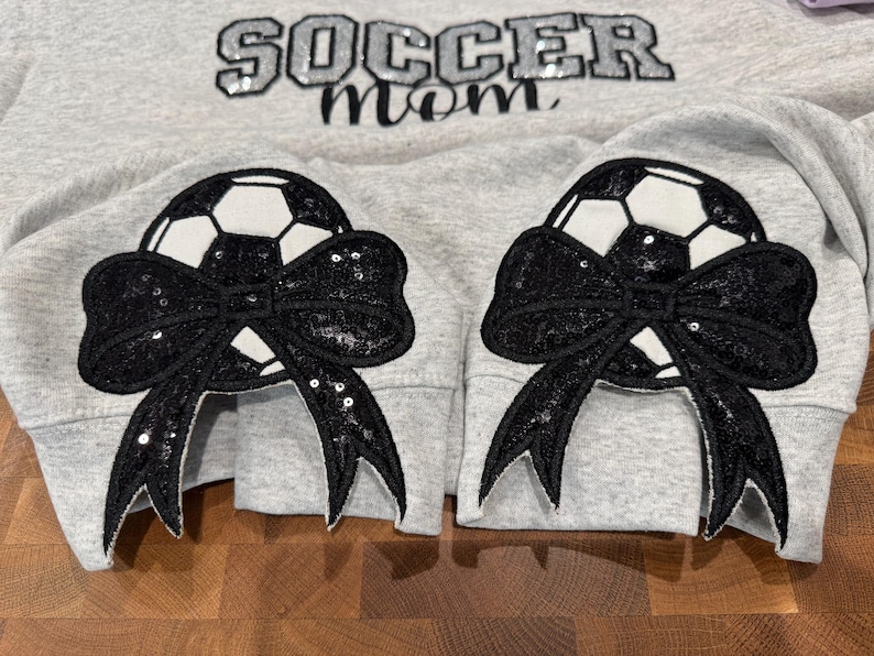 May include: A light gray sweatshirt featuring the text SOCCER MOM in black sequined letters. Each sleeve cuff displays a large black sequined bow layered over a white and black soccer ball graphic, creating a decorative detail on the garment.