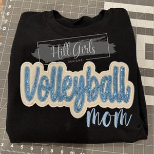 Puede incluir: Una sudadera negra de cuello redondo con un gran parche aplicado con la palabra Volleyball en lentejuelas azul claro sobre un fondo crema. Debajo del parche, la palabra mom est&aacute; bordada en azul claro. Un logo dice Hill Girls DESIGNS.