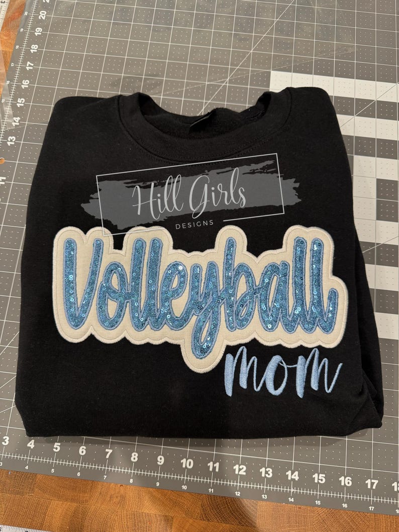 Puede incluir: Una sudadera negra de cuello redondo con un gran parche aplicado con la palabra Volleyball en lentejuelas azul claro sobre un fondo crema. Debajo del parche, la palabra mom est&aacute; bordada en azul claro. Un logo dice Hill Girls DESIGNS.
