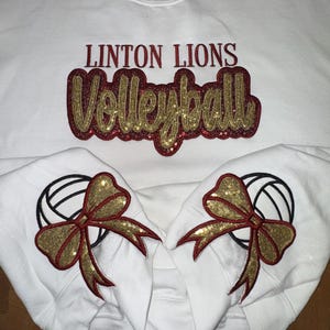Puede incluir: Una sudadera blanca de cuello redondo con el texto LINTON LIONS en rojo oscuro y la palabra Volleyball en letra cursiva de purpurina dorada con borde rojo oscuro. Las mangas muestran balones de voleibol bordados con lazos dorados.