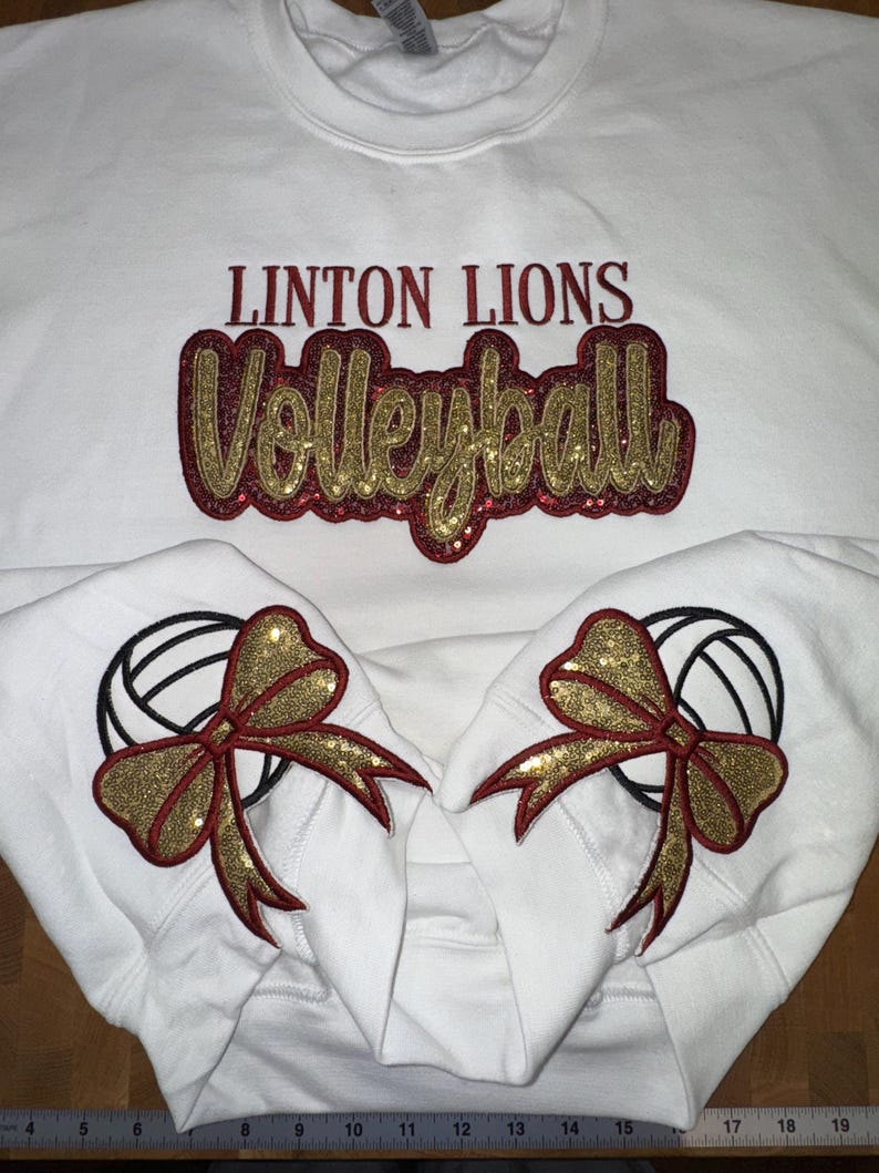 Puede incluir: Una sudadera blanca de cuello redondo con el texto LINTON LIONS en rojo oscuro y la palabra Volleyball en letra cursiva de purpurina dorada con borde rojo oscuro. Las mangas muestran balones de voleibol bordados con lazos dorados.