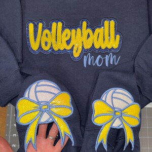 May include: Navy blue sweatshirt with the words "Volleyball mom" in yellow and light blue. The sleeves feature a volleyball with a yellow bow. A hand is holding the sleeve.