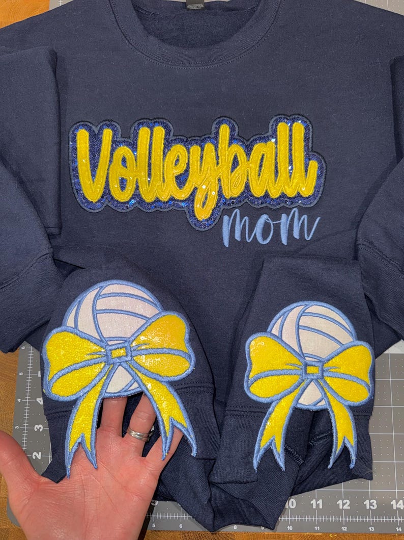 Puede incluir: Una sudadera azul marino con el texto Volleyball mom en letras brillantes amarillas y azules. Las mangas muestran parches de un voleibol blanco con un gran lazo amarillo brillante. Se observa una regla con medidas en cent&iacute;metros.