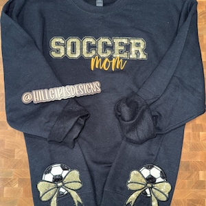 May include: A black long-sleeve sweatshirt featuring the text SOCCER in gold glitter block letters and mom in gold script. Each sleeve cuff displays a black and white soccer ball graphic adorned with a large, shimmering gold glitter bow.