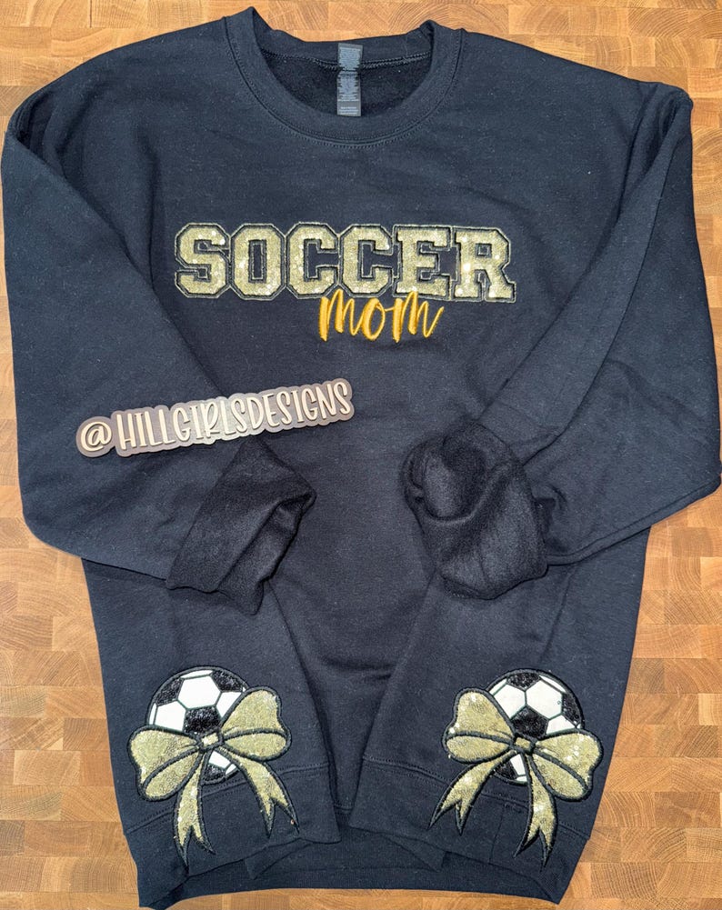 May include: A black long-sleeve sweatshirt featuring the text SOCCER in gold glitter block letters and mom in gold script. Each sleeve cuff displays a black and white soccer ball graphic adorned with a large, shimmering gold glitter bow.
