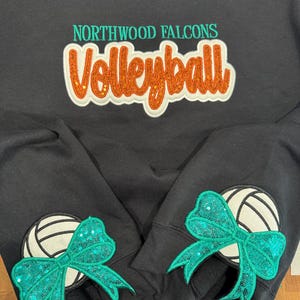 Puede incluir: Una sudadera negra de cuello redondo con el texto NORTHWOOD FALCONS en color verde azulado y la palabra Volleyball en letras naranjas con purpurina. Cada manga muestra un gr&aacute;fico de voleibol blanco con un gran lazo de lentejuelas verde azulado.