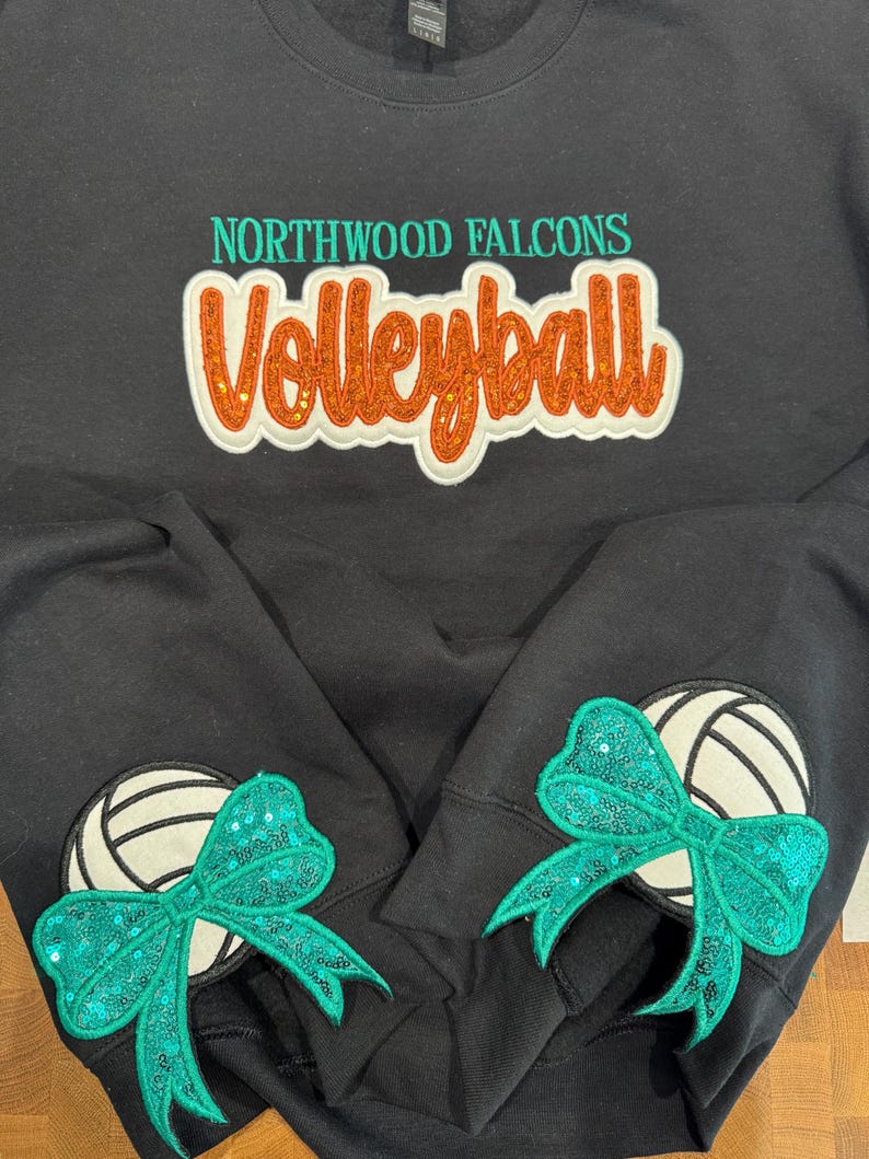 Puede incluir: Una sudadera negra de cuello redondo con el texto NORTHWOOD FALCONS en color verde azulado y la palabra Volleyball en letras naranjas con purpurina. Cada manga muestra un gr&aacute;fico de voleibol blanco con un gran lazo de lentejuelas verde azulado.