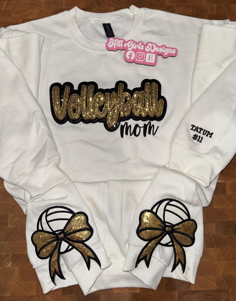 Puede incluir: Una sudadera blanca de manga larga con la palabra Volleyball en purpurina dorada con borde negro, y la palabra mom en letra cursiva negra. Las mangas muestran lazos de purpurina dorada con gr&aacute;ficos de voleibol y el texto TATUM #11.