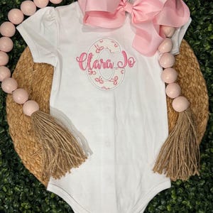May include: White baby bodysuit with short sleeves, featuring a pink bow and the embroidered name "Clara Jo" inside a floral-patterned letter "C". The bodysuit is displayed on a woven mat with decorative pink beads and tassels.