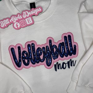 Puede incluir: Una sudadera blanca de manga larga con la palabra Volleyball en letras de lentejuelas azul oscuro con un borde rosa, y la palabra mom en letra cursiva azul oscuro debajo. Una etiqueta rosa con el texto Hill Girls Designs est&aacute; adjunta.
