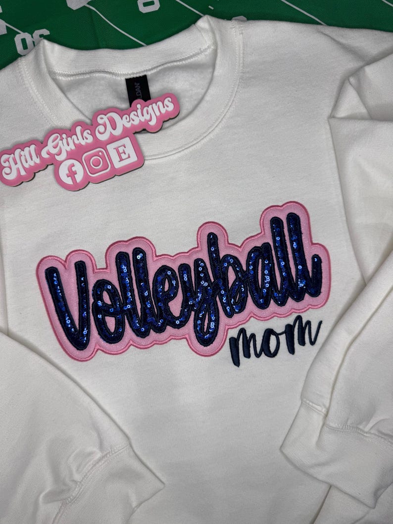 Puede incluir: Una sudadera blanca de manga larga con la palabra Volleyball en letras de lentejuelas azul oscuro con un borde rosa, y la palabra mom en letra cursiva azul oscuro debajo. Una etiqueta rosa con el texto Hill Girls Designs est&aacute; adjunta.