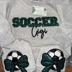 May include: A light gray crewneck sweatshirt featuring the word SOCCER in dark green glittery block letters with the word Gigi in black script below. Each sleeve displays a black and white soccer ball graphic accented with a dark green glitter bow.