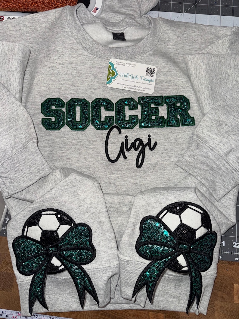 May include: A light gray crewneck sweatshirt featuring the word SOCCER in dark green glittery block letters with the word Gigi in black script below. Each sleeve displays a black and white soccer ball graphic accented with a dark green glitter bow.