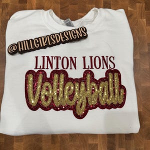 Puede incluir: Una sudadera blanca de cuello redondo con el texto LINTON LIONS en letras de color rojo oscuro sobre la palabra Volleyball, escrita en cursiva dorada con un fondo de lentejuelas de color rojo oscuro. Un letrero de madera dice @HILLGIRLSDESIGNS.