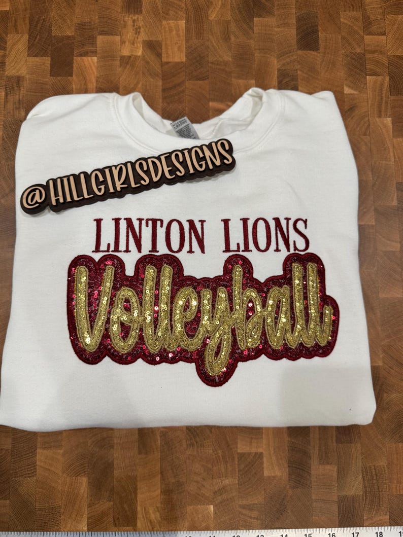 Puede incluir: Una sudadera blanca de cuello redondo con el texto LINTON LIONS en letras de color rojo oscuro sobre la palabra Volleyball, escrita en cursiva dorada con un fondo de lentejuelas de color rojo oscuro. Un letrero de madera dice @HILLGIRLSDESIGNS.