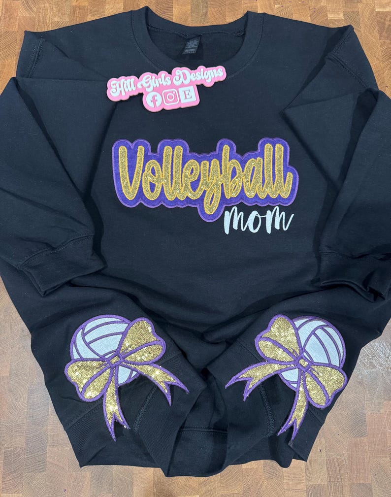 Puede incluir: Una sudadera negra de manga larga con el texto Volleyball mom en purpurina dorada y letra morada. Dos gr&aacute;ficos de voleibol con lazos de purpurina dorada est&aacute;n en las mangas. Un logotipo rosa de Hill Girls Designs se encuentra en la parte superior.