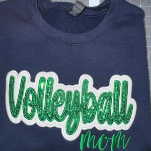 Puede incluir: Una sudadera azul marino de cuello redondo que presenta la palabra Volleyball en letras grandes de lentejuelas verdes con un borde de fieltro blanco, situada sobre la palabra mom en letras cursivas bordadas en verde en el frente.