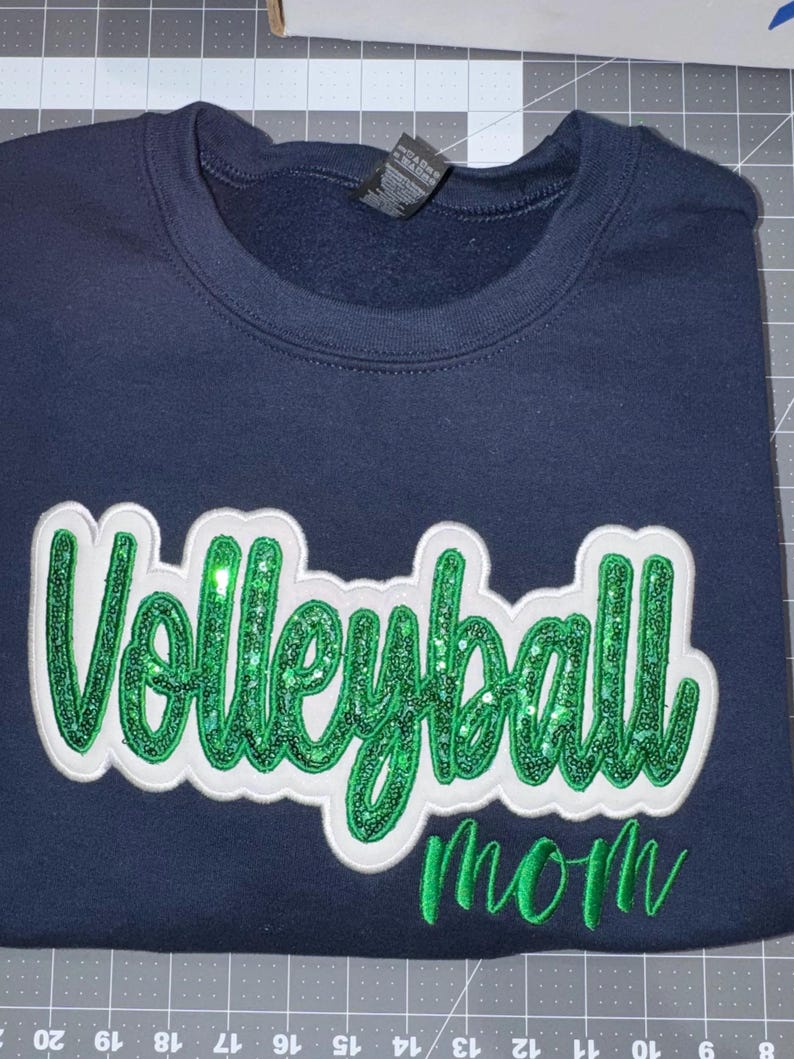 Puede incluir: Una sudadera azul marino de cuello redondo que presenta la palabra Volleyball en letras grandes de lentejuelas verdes con un borde de fieltro blanco, situada sobre la palabra mom en letras cursivas bordadas en verde en el frente.