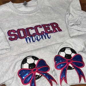 May include: A light gray heathered sweatshirt featuring the text SOCCER mom embroidered in red and blue glittery thread. The sleeves display two soccer ball patches, each adorned with a large, sparkling blue sequined bow with red trim.