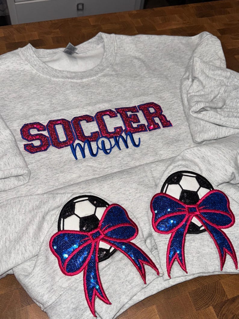 May include: A light gray heathered sweatshirt featuring the text SOCCER mom embroidered in red and blue glittery thread. The sleeves display two soccer ball patches, each adorned with a large, sparkling blue sequined bow with red trim.