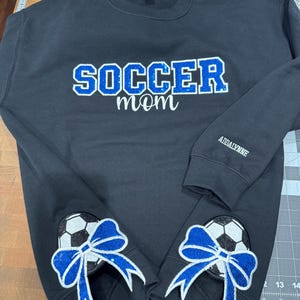 May include: A black crewneck sweatshirt featuring the word SOCCER in blue glitter letters with the word mom in white script below. The left sleeve has the name ADDALYNNE in white, and the bottom hem displays two soccer ball patches with blue glitter bows.