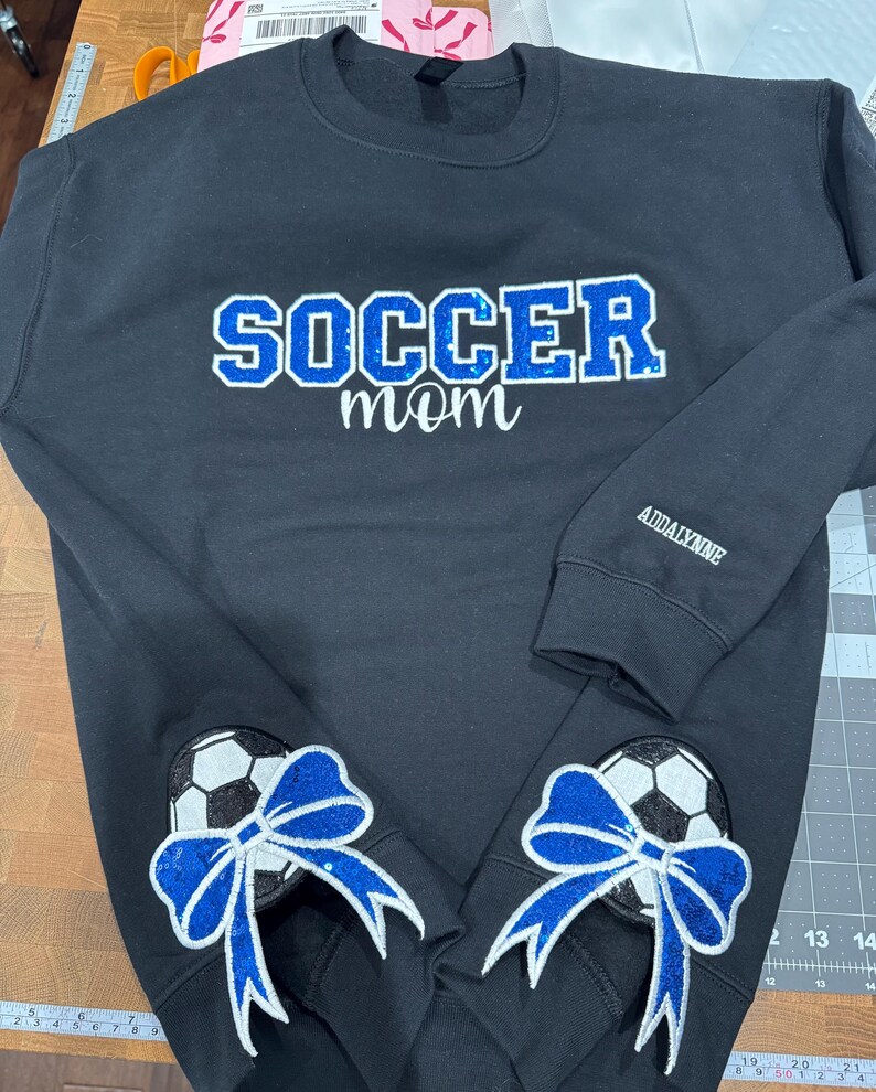 May include: A black crewneck sweatshirt featuring the word SOCCER in blue glitter letters with the word mom in white script below. The left sleeve has the name ADDALYNNE in white, and the bottom hem displays two soccer ball patches with blue glitter bows.