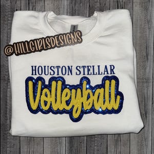 Puede incluir: Una sudadera blanca de cuello redondo con el texto HOUSTON STELLAR en letras de bloque azul oscuro sobre la palabra Volleyball en cursiva amarilla. La palabra Volleyball est&aacute; superpuesta sobre un parche azul oscuro con purpurina.