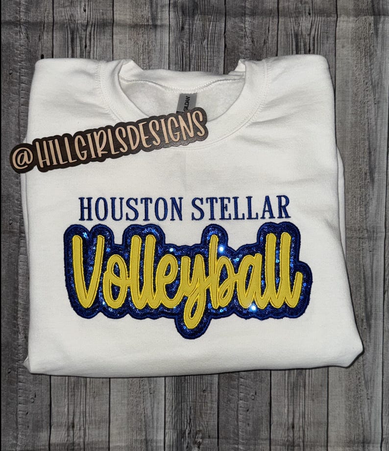 Puede incluir: Una sudadera blanca de cuello redondo con el texto HOUSTON STELLAR en letras de bloque azul oscuro sobre la palabra Volleyball en cursiva amarilla. La palabra Volleyball est&aacute; superpuesta sobre un parche azul oscuro con purpurina.