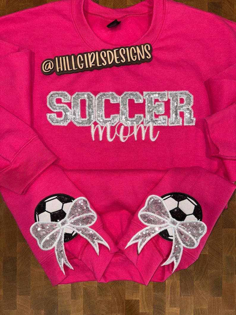 May include: A bright pink long-sleeve sweatshirt featuring the text @HILLGIRLSDESIGNS in black, with the word SOCCER in silver glitter block letters and mom in white script. Each sleeve cuff displays a black and white soccer ball with a silver sequin bow.