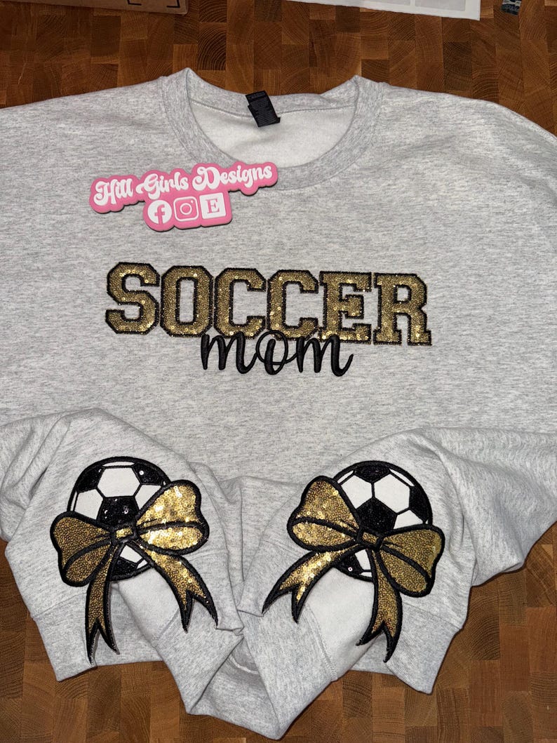 May include: A light gray crewneck sweatshirt featuring the text "SOCCER mom" in gold glitter and black script lettering. Each sleeve cuff displays a black and white soccer ball graphic adorned with a large, gold glitter sequin bow.
