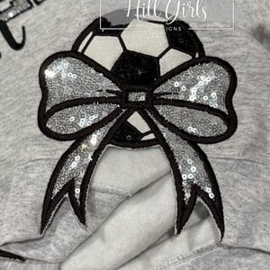 May include: A light gray sweatshirt featuring a large applique of a soccer ball adorned with a prominent silver sequined bow. The soccer ball has black and white panels with glittery accents. The text Hill Girls DESIGNS is visible above.