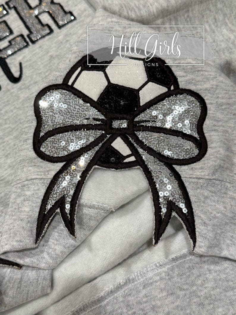 May include: A light gray sweatshirt featuring a large applique of a soccer ball adorned with a prominent silver sequined bow. The soccer ball has black and white panels with glittery accents. The text Hill Girls DESIGNS is visible above.