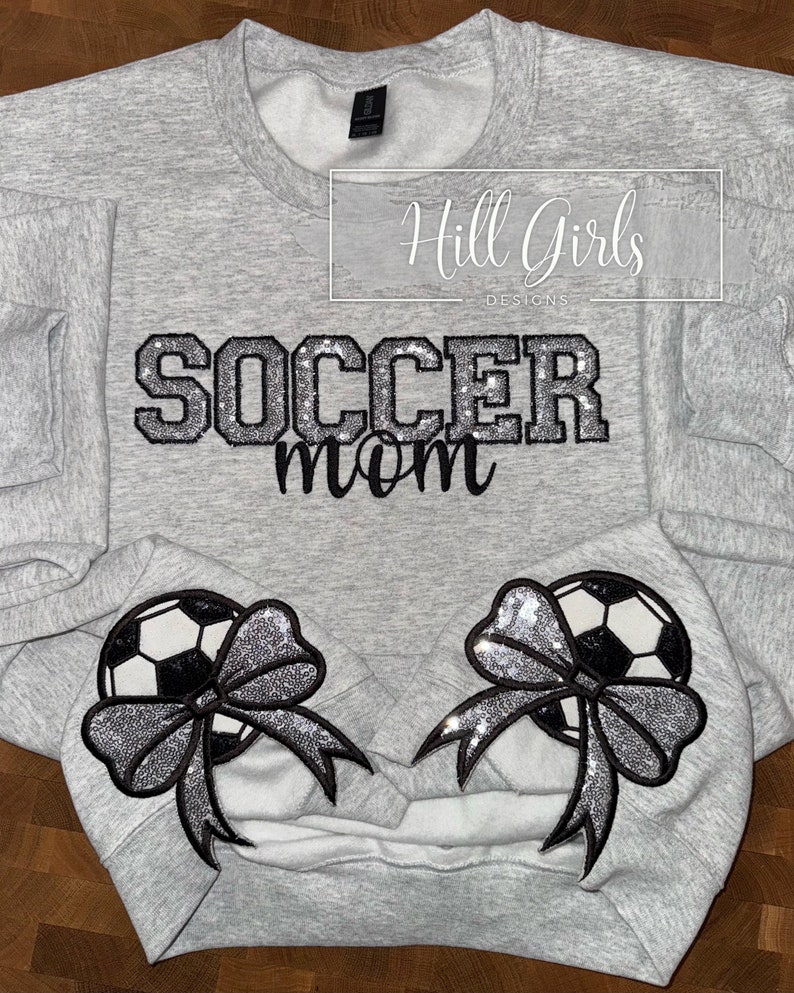 May include: A light gray crewneck sweatshirt featuring the text SOCCER mom in black and silver glitter lettering. Two embroidered patches are positioned at the bottom, each depicting a black and white soccer ball adorned with a silver glitter bow.