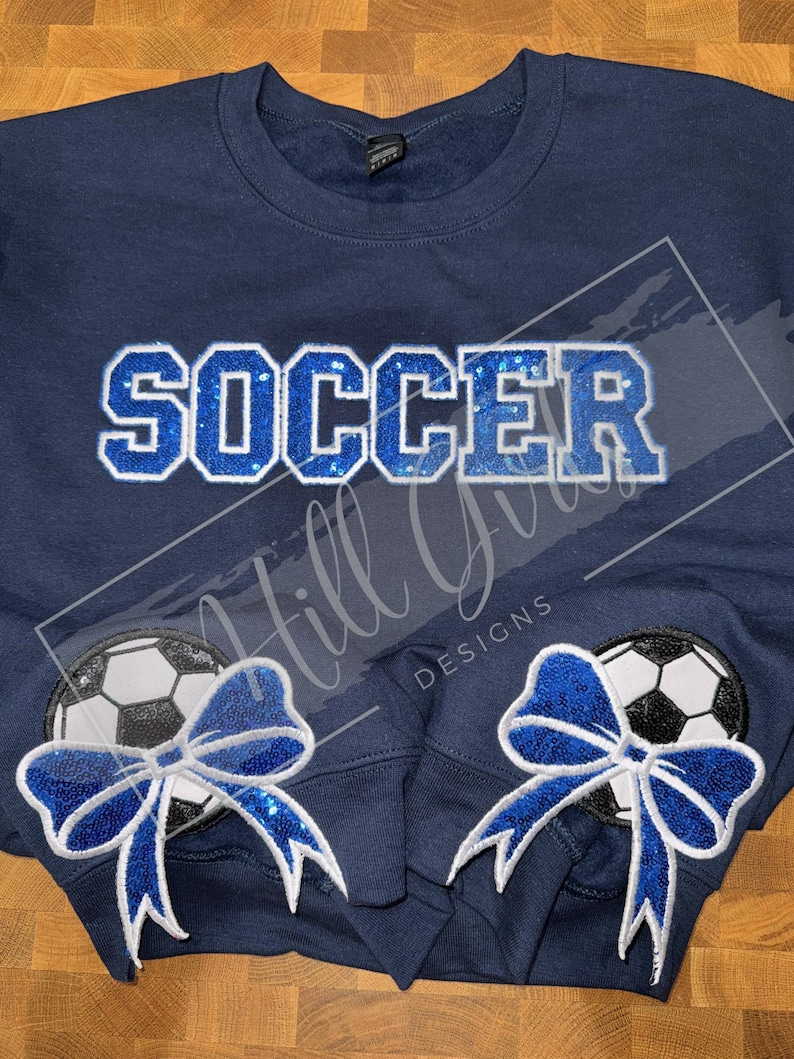 May include: A navy blue crewneck sweatshirt featuring the word SOCCER in blue sequined block letters across the chest. The sleeves display circular patches of black and white soccer balls adorned with large, bright blue sequined bows.