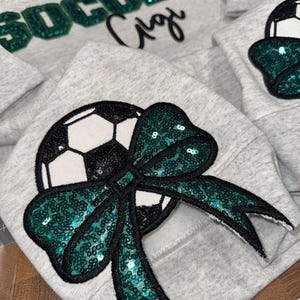 May include: A light gray sweatshirt featuring a large sequined soccer ball graphic adorned with a dark green sequined bow. The word SOCCER is written in dark green sequins above the script text Gigi. A measuring tape shows 50 cm.