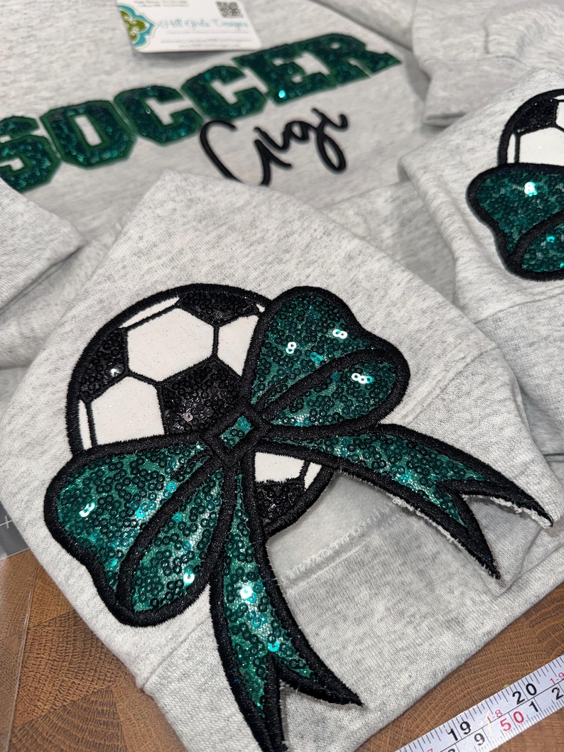 May include: A light gray sweatshirt featuring a large sequined soccer ball graphic adorned with a dark green sequined bow. The word SOCCER is written in dark green sequins above the script text Gigi. A measuring tape shows 50 cm.