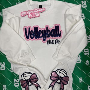 Puede incluir: Una sudadera blanca de manga larga con el texto "Volleyball mom" en letras azul oscuro con purpurina y borde rosa. Dos gr&aacute;ficos de voleibol con lazos rosas de purpurina est&aacute;n situados en los pu&ntilde;os inferiores de las mangas.