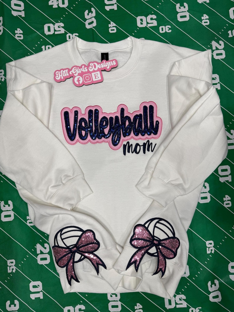Puede incluir: Una sudadera blanca de manga larga con el texto "Volleyball mom" en letras azul oscuro con purpurina y borde rosa. Dos gr&aacute;ficos de voleibol con lazos rosas de purpurina est&aacute;n situados en los pu&ntilde;os inferiores de las mangas.