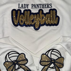 Puede incluir: Una sudadera blanca con el texto LADY PANTHERS en azul oscuro y la palabra Volleyball en letra cursiva con purpurina dorada y borde de lentejuelas azul oscuro. Cada pu&ntilde;o muestra un gr&aacute;fico de voleibol con un lazo de lentejuelas doradas.