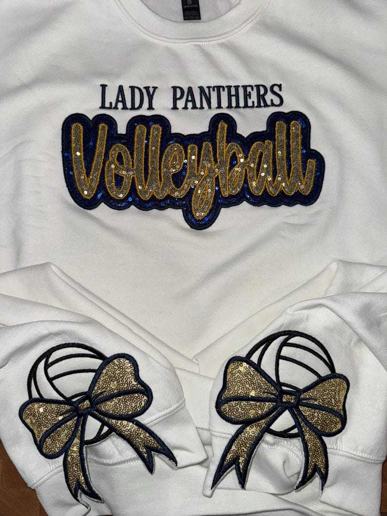 Puede incluir: Una sudadera blanca con el texto LADY PANTHERS en azul oscuro y la palabra Volleyball en letra cursiva con purpurina dorada y borde de lentejuelas azul oscuro. Cada pu&ntilde;o muestra un gr&aacute;fico de voleibol con un lazo de lentejuelas doradas.