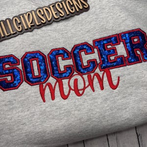 May include: A light gray sweatshirt featuring the text "SOCCER mom" embroidered on the front. The word "SOCCER" is in block letters filled with blue sequins and outlined in red thread, while "mom" is in red cursive script. A logo reads "LLGIRLSDESIGNS".