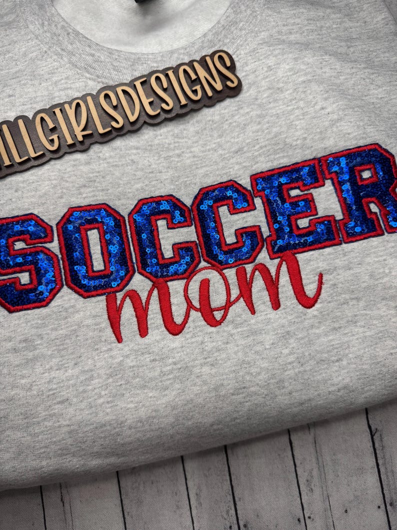 May include: A light gray sweatshirt featuring the text "SOCCER mom" embroidered on the front. The word "SOCCER" is in block letters filled with blue sequins and outlined in red thread, while "mom" is in red cursive script. A logo reads "LLGIRLSDESIGNS".