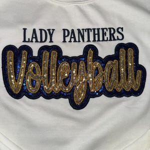 Puede incluir: Una sudadera blanca de cuello redondo con el texto LADY PANTHERS en letras de bloque azul oscuro sobre la palabra Volleyball en letra cursiva grande. Las letras cursivas est&aacute;n rellenas de lentejuelas doradas y bordeadas en tela azul oscuro.