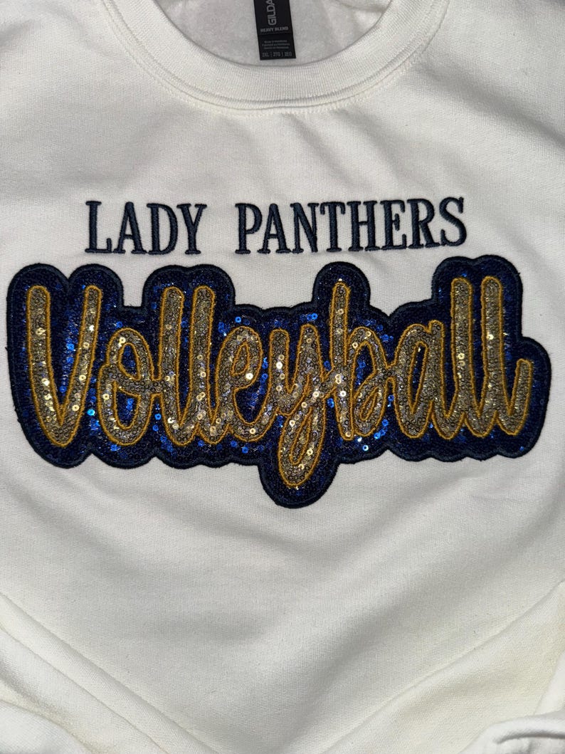 Puede incluir: Una sudadera blanca de cuello redondo con el texto LADY PANTHERS en letras de bloque azul oscuro sobre la palabra Volleyball en letra cursiva grande. Las letras cursivas est&aacute;n rellenas de lentejuelas doradas y bordeadas en tela azul oscuro.