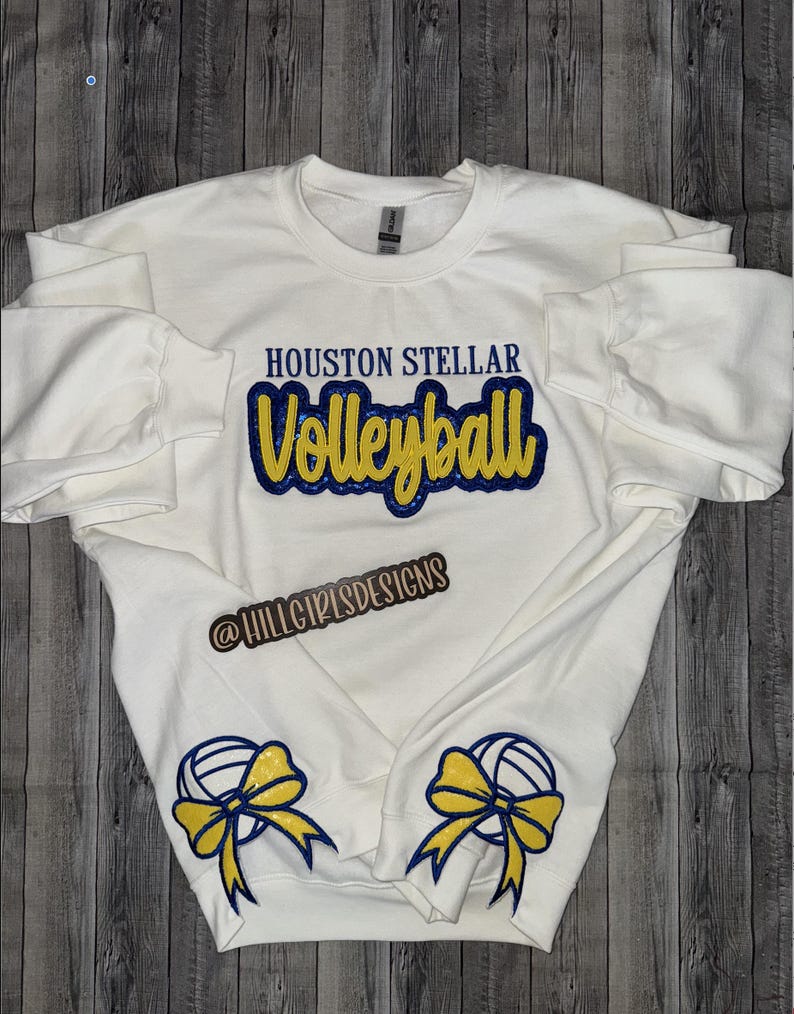 May include: White sweatshirt with the text "HOUSTON STELLAR Volleyball" in blue and yellow lettering. The text "@HILLGIRLSDESIGNS" is below the main text. The sleeves have a yellow bow with a blue volleyball on each cuff.