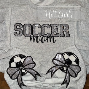 May include: A light gray crewneck sweatshirt featuring the text SOCCER mom in black and silver glitter lettering. Two black and white soccer ball graphics, each adorned with a large silver sequin bow, are positioned on the lower front section.