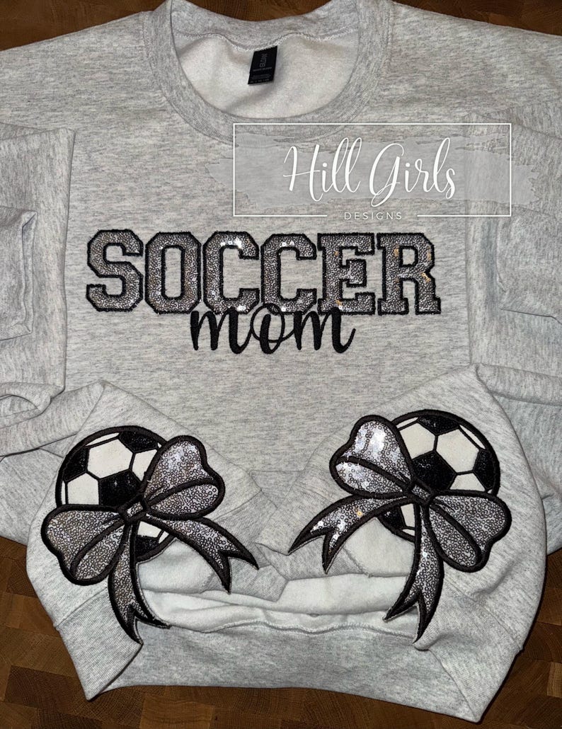 May include: A light gray crewneck sweatshirt featuring the text SOCCER mom in black and silver glitter lettering. Two black and white soccer ball graphics, each adorned with a large silver sequin bow, are positioned on the lower front section.