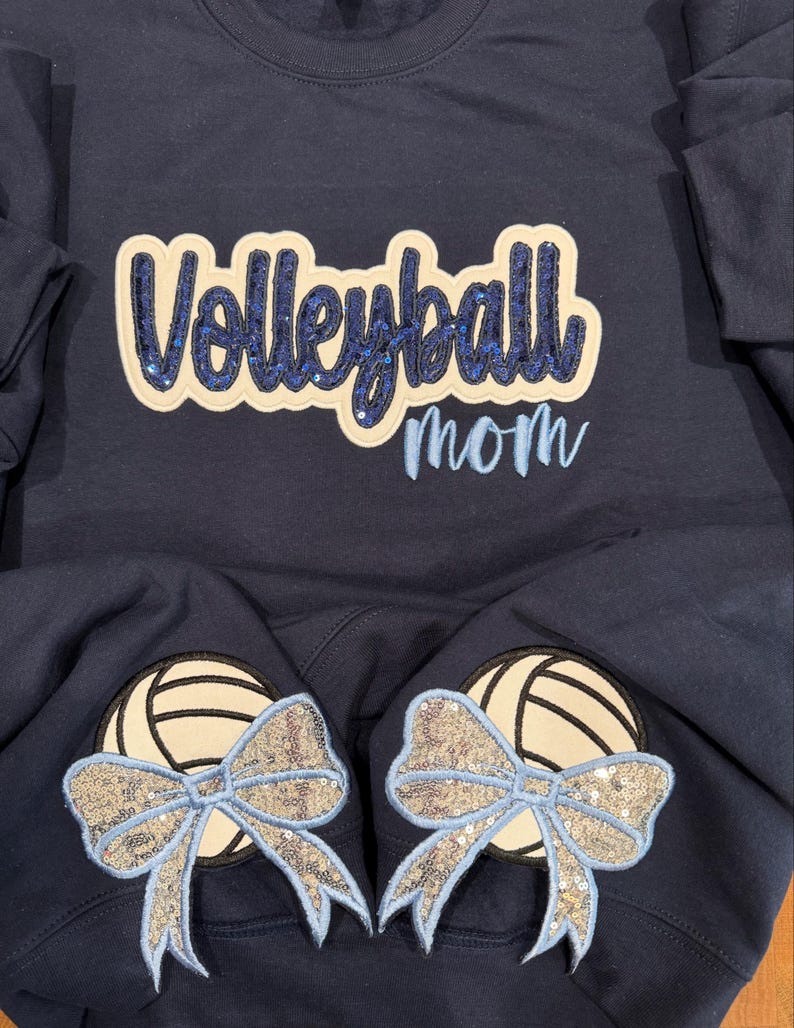 Puede incluir: Una sudadera azul marino con la palabra Volleyball en letras de lentejuelas azul oscuro sobre un parche crema, con la palabra mom bordada en azul claro debajo. Dos parches de voleibol crema con lazos de lentejuelas azul claro en las mangas.